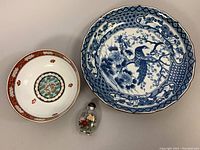 Photo showing three Asianware pieces including large blue and white porcelain bowl, smaller decorative bowl with red motif, and small glass snuff bottle