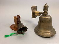 Photo of the bronze butler's bell mounted on wood and the brass nautical wall bell together on a neutral background.