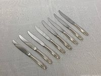 Full set of 8 Wallace dinner knives arranged side by side on a white patterned cloth, clearly showing the sterling silver detail on the handles and stainless steel blades.
