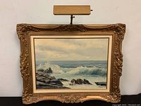 Front view of the signed oil painting showing ocean waves and rocks inside carved gold ornate frame with picture light mounted on top.