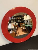 Front view of the red framed round mirror leaning against a wall in a room with other furniture.