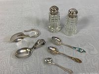 Overview shot showing two cut crystal shakers with sterling silver lids plus five assorted sterling silver spoons including floral enamel handle spoon and sugar tongs.