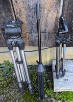 Three camera tripods leaning against a wooden surface outdoors, showing different models and brands including Platinum Plus by Sunpak and SLIK