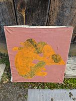 Full view of abstract painting on pink canvas with orange and green figures, edges of canvas visible showing wear and dirt on surroundings.