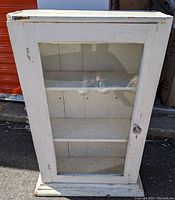 Full front view of white cabinet with glass door and crystal knob, showing three shelves inside.