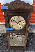 Full view of the antique wooden wall clock showing clock face and lower mirrored panel with decorative metalwork.