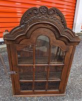 Front view of small antique wooden cabinet with glass door and carved decorative top.