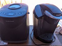Black Hamilton Beach bread maker and black Keurig coffee machine sitting side-by-side on a table.