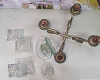 Four candlesticks including three metal with red decorative accents arranged top right and one clear glass candlestick with geometric base bottom left, all placed on a white surface beneath a colorful painting.