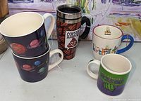 Five assorted coffee cups and mugs displayed on a surface, showing designs and styles of each.