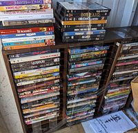 Photo of three tall stacks of DVDs vertically arranged on a wooden shelving unit, showing a variety of DVD titles including animated movies and adult films.