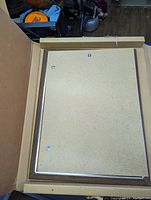 Photo showing the assembled wooden frame with backing board secured by metal clips inside a cardboard box.