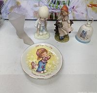 Photo showing all five items: Precious Moments boy figurine with crutches, seated girl figurine, Holly Hobby plate, small bell, and frosted glass vase.