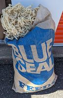 Large burlap feed sack with 'Blue Seal Feeds' logo, filled with thick off-white wool yarn visible inside sack opening.
