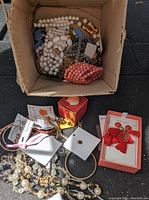 Box containing various beaded necklaces and bracelets, strands of white, coral, and gray beads, alongside miscellaneous chain jewelry.