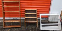 Three wood shelves displayed in front of an orange door: one large natural wood shelf, one medium natural wood shelf, and one small white painted shelf.