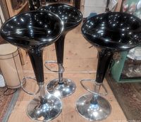 Three black and silver modern bar stools seen from front angle showing black molded seats and chrome bases with footrests.