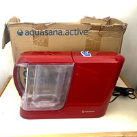 Front and side view of Aquasana Clean Water Machine with transparent water reservoir attached, original box behind showing branding.