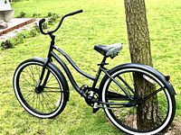 Full side view of black beach cruiser bicycle with 26 inch wheels and whitewall tires parked on grass near tree.
