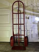 Front view of red two-wheel hand truck dolly with metal frame, two black rubber wheels, vertical handle and flat loading plate shown against a garage door background.