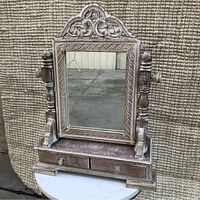 Front view of wooden mirror with decorative carvings and two small drawers beneath the mirror. Mirror glass is rectangular and frame shows a distressed finish.
