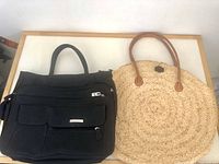 Two purses placed side by side on a wooden surface. One is black fabric with zippers, the other woven straw purse.