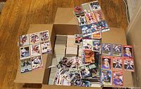 Photo showing a large open box filled with stacked sports cards including baseball cards, some cards laid out on top, with plastic protective sleeves and rookie/gold stickers on sleeves.