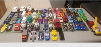 Photo showing assortment of toy cars including race cars, trucks, and unique designs like a dinosaur car, all arranged in rows.