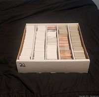 Front-facing image of a cardboard box divided into 5 compartments filled with large stacks of trading cards in plastic protectors, showing primarily the back sides of cards which are mostly white with some color variation.