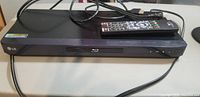 Front view of the black LG Blu-Ray and DVD player showing the disc tray and control buttons. Remote control placed on top along with power cord and HDMI cable.