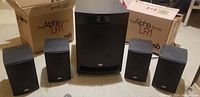 Four PSB Alpha Series LR1 bookshelf speakers with two original boxes and one PSB SubSeries 125 powered subwoofer in front.