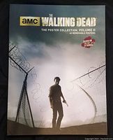 Front cover of the poster book featuring a character from The Walking Dead near barbed wire fencing with title and logo.