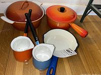 Overview of the set showing the large orange casserole dish with lid, orange pot with lid, blue-handled small skillet, and the two ladles (orange and blue enamel bodies with black handles)