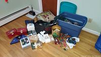 Wide shot showing assortment of boxed Christmas ornaments, decorative bags, a blue plastic storage bin, and other Christmas décor on the floor.
