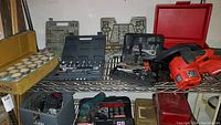 Photo shows a variety of hand tools and hardware organized in multiple plastic cases and toolboxes, including socket sets, drill bits, screws, and wrenches, with some power tools on lower shelves.