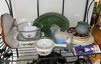 Photo shows multiple Corningware casserole dishes and bowls, green ceramic plate, various ceramic plates, and Henckels garlic press box on shelf