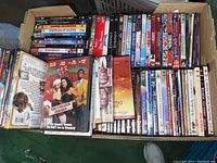 Top-down photo showing a large collection of mixed genre DVDs in a cardboard box with visible titles such as Casino Royale and Sleepless in Seattle.