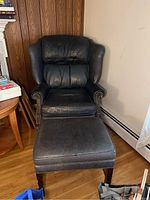 Front view showing black leather wingback armchair with matching ottoman in front on wood floor next to wood panel wall.