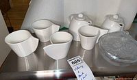 Four white porcelain cups, two jugs and clear glass bowl arranged on stainless surface