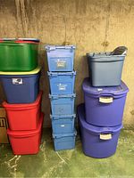 Twelve plastic storage tubes stacked and arranged in groups by color and type against a basement concrete wall.