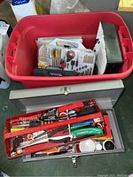 Full view of red plastic bin filled with a drill bit set, a black rectangular metal box, and other assorted hardware items, next to a gray metal toolbox with red tray insert showing various hand tools.