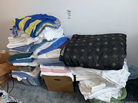 Stack of assorted bath towels in various colors with a pile of white linens and a dark patterned comforter on top.