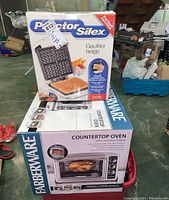 Boxes of Proctor Silex waffle iron and Farberware countertop oven stacked in a basement setting.