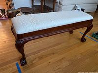 Full view of wooden claw foot bench with beige upholstered cushion showing the entire bench and cushion.