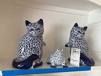 Two matching ceramic cats and one frog in blue and white ceramic on display shelf with auction tag