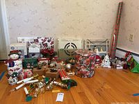 Wide shot of Christmas decor assortment showing snowmen figures, tins, electric candles, gift wrap, and wall decor arranged on hardwood floor against wallpaper background.