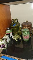 Full view of the set showing a large green frog cookie jar, a smaller frog planter or sponge holder, a brown cookie canister, a peach-themed pitcher, and three small square canisters with fruit designs.