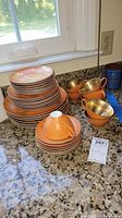 Full display of stacked orange and gold Dorothy Thorpe dishware including cups, plates, and saucers on countertop with granite pattern.