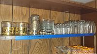 Wooden shelf holding various glasses, some decorative with gold details, others clear, arranged in rows.