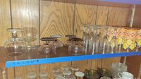 Set of various glassware items on a wooden shelf including clear stemmed glasses, smaller rounded stemmed glasses, and decorative glasses with floral patterns. Some mildew visible.
